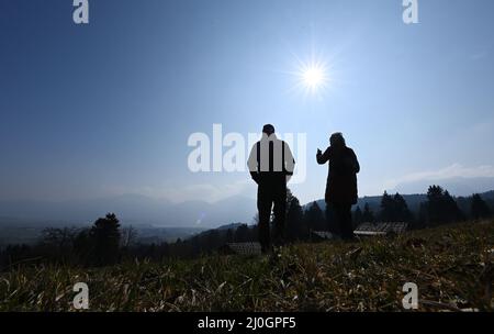 19 marzo 2022, Baviera, Großweil: Due visitatori camminano tra le storiche capanne alpine sui terreni del Museo all'aperto Glentleiten attraverso un prato. Dopo la polvere del Sahara, il sole primaverile si ripresenta nel cielo nuvoloso. Foto: Angelika Warmuth//dpa Foto Stock