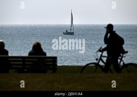 Langenargen, Germania. 19th Mar 2022. Una barca naviga sul lago di Costanza al largo di Langenargen, mentre in primo piano le persone si siedono su una panchina nel parco sul lago e un ciclista spinge la sua bicicletta oltre. Credit: Felix Kästle/dpa/Alamy Live News Foto Stock