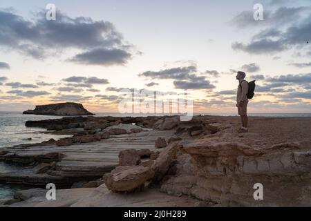 L'uomo con zaino sta camminando lungo una costa rocciosa al tramonto vicino al mare Mediterraneo a Cipro. Maschio turistico ammira la vista Ye Foto Stock