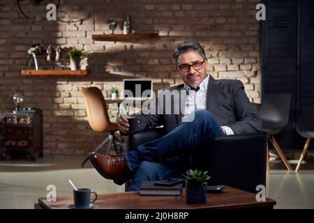 Uomo d'affari seduto in poltrona in un accogliente ufficio domestico, sorridente, bere caffè. Foto Stock