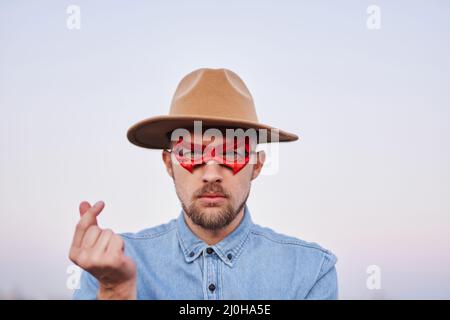 Carino uomo caucasico bearded in rosso maschera di supereroe e maglietta jeans in posa con il volto serio davanti alla macchina fotografica. Maschio che mostra cuore coreano o mini cuore gesto all'aperto al tramonto con vista urbana Foto Stock