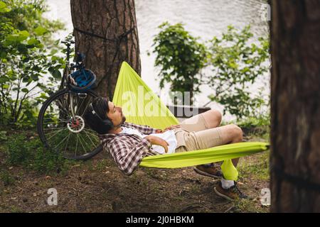 L'uomo viaggia in bicicletta, rilassandosi in amaca verde, navigando su Internet su smartphone, ascoltando musica sulle cuffie nella foresta vicina Foto Stock