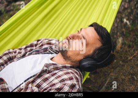 Giovane uomo caucasico che riposa in amaca, ascoltando musica con le cuffie e utilizzando smartphone in seguito in bicicletta in foresta ne Foto Stock