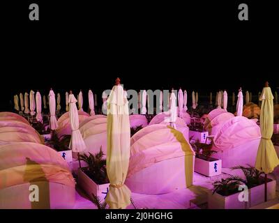 Divani spiaggia semicircolari con baldacchino e ombrelloni tra loro si trovano in un'area ricreativa sulla riva del mare di notte in rosa li Foto Stock