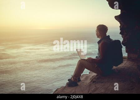 Connessione con il mondo. Scatto a tutta lunghezza di un giovane uomo che usa il suo computer portatile mentre si siede su una scogliera in montagna. Foto Stock