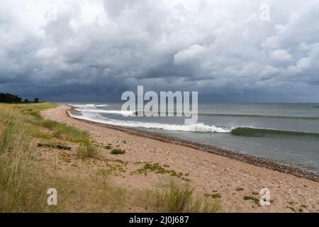 Serie di onde pulite che si infrangono su una spiaggia di sabbia con erba palude e dune di sabbia e un cielo oscuro e ominoso Foto Stock