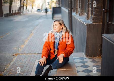 Felice sorridente donna di mezza età in giacca arancione giù seduta su scale di cemento all'aperto Foto Stock