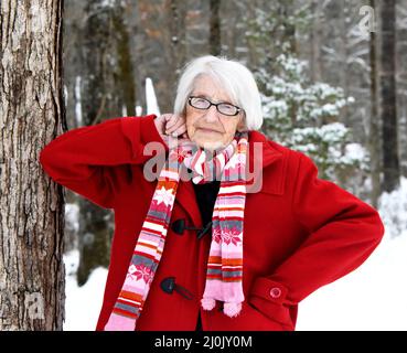 Donna anziana, di 100 anni, appoggia il gomito contro un albero. È impacchettata in un cappotto rosso brillante e sciarpa a righe. La scena invernale la circonda. Foto Stock