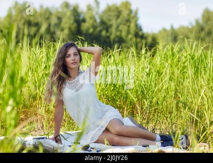 Giovane donna bionda in abito bianco è seduta su un foglio da picnic in erba alta. Foto Stock
