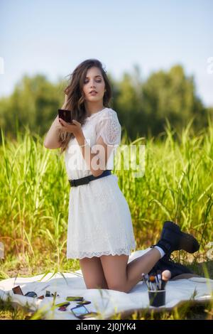 Giovane donna bionda in abito bianco è inginocchiato su un foglio da picnic in erba alta. Foto Stock