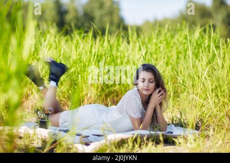 Giovane donna bionda in abito bianco si trova su un foglio da picnic in erba alta. Foto Stock