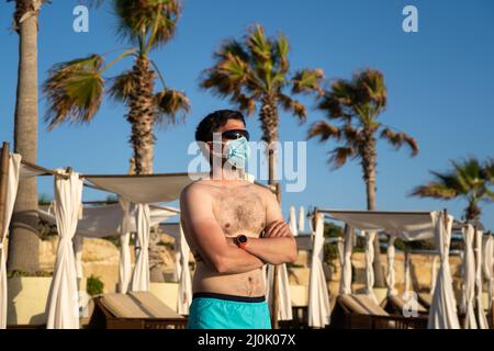 L'uomo in maschera medica si erge da solo su una spiaggia vuota vicino a lettini e palme a Cipro, città di Paphos durante il coronavirus quara Foto Stock