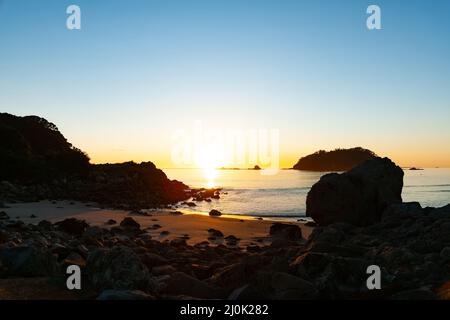 In silhouette il litorale roccioso e l'isola sono solo essere illuminati da caldo sole mattutino su un orizzonte lontano. Foto Stock