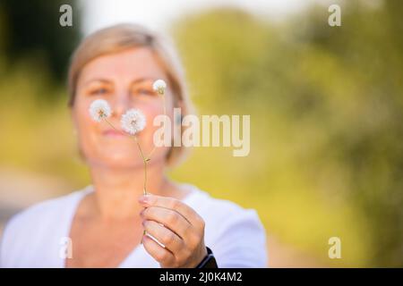 Primo piano di una mano donna con manicure rosa e orologio digitale che tiene in mano il fiore di dente di leone con la donna bionda sullo sfondo. Foto Stock