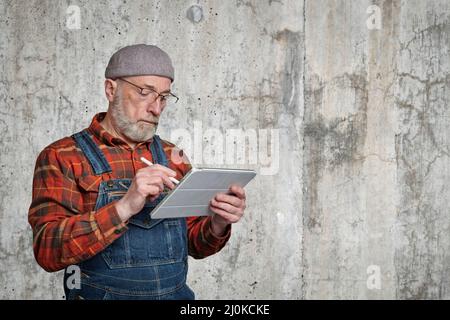 un uomo anziano sicuro che indossa occhiali, una camicia in flanella e tute sta prendendo appunti o schizzi su un tablet digitale Foto Stock