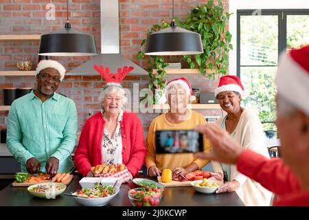 Gruppo di amici anziani e donne felice diversi in cappelli di natale prendendo selfie in cucina Foto Stock