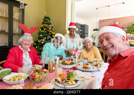 Gruppo vario di amici anziani felici in cappelli di festa che celebrano il natale insieme, prendendo selfie Foto Stock