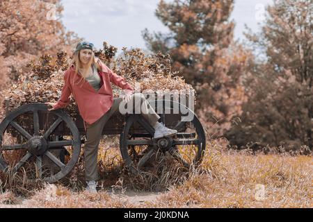 Una ragazza ridente in un abito casual si alza appoggiando il piede e la mano sulle ruote di un carrello di legno in un parco autunnale. Foto Stock