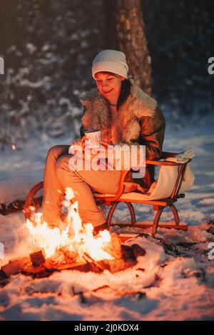Felice giovane donna in caldo cappello a maglia e giacca siede su slitta di legno da falò con tazza di tè caldo Foto Stock
