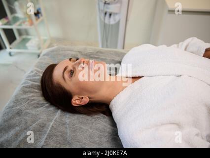 Giovane bella donna caucasica sdraiata su un lettino da massaggio in una clinica cosmetologica, rilassante durante il check-up medico, pronta a ricevere un anti-invecchiamento, Foto Stock