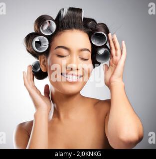 Infilando i ricci. Studio girato di una giovane donna attraente in posa con arricciatori nei suoi capelli su uno sfondo grigio. Foto Stock