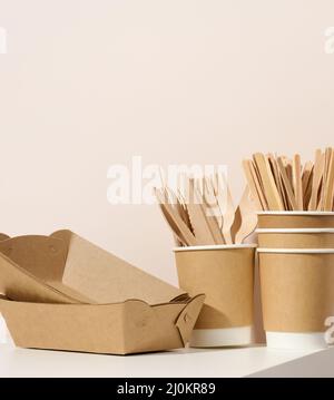 Lastre e tazze di cartone marrone, forcelle e coltelli di legno su un tavolo bianco, sfondo beige. Stoviglie ecocompatibili Foto Stock