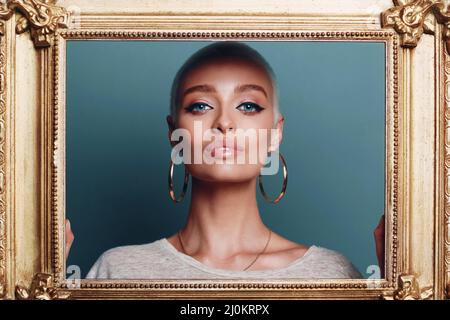 Giovane donna millenaria con capelli biondi corti tiene la cornice dorata in mani dietro il suo ritratto del viso. Foto Stock