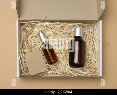 Bottiglie di vetro marrone per cosmetici e cartellini di carta su una corda giacciono in una scatola di carta di cartone su sfondo beige, vista dall'alto Foto Stock