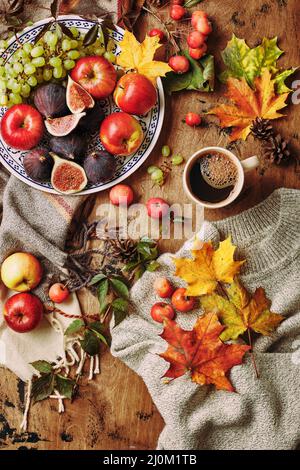 Piatto con fichi, mele e uva e tazza di caffè su sfondo ligneo con un caldo maglione, sciarpa, foglie d'autunno e mele. R Foto Stock