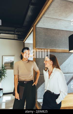 Donne d'affari di successo che condividono una risata in ufficio. Due giovani imprenditori femminili ridono allegro mentre si levano in piedi insieme in un workspa moderno Foto Stock