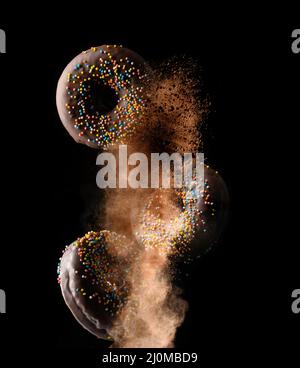 Ciambelle rotonde al cioccolato con zucchero multicolore cosparge di levitate in una nuvola di cacao marrone su sfondo nero Foto Stock