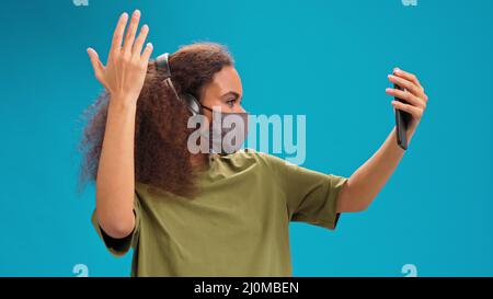 Realizzazione di un video ragazza afroamericana utilizzando il suo smartphone con cuffie in t-shirt a oliva, maschera facciale riutilizzabile, per prevenire coron Foto Stock