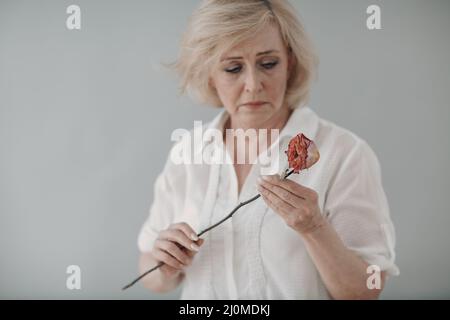 Donna anziana sconvolta in bianco vestita tenere appassito un vecchio fiore di rosa secco. Foto Stock