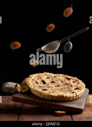 Torta rotonda al forno con prugne su un piano di cottura in legno, setaccio e fettine di frutta prugna su sfondo nero Foto Stock