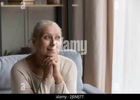 La donna più anziana si siede sul divano, sorridendo fissando fuori la finestra Foto Stock