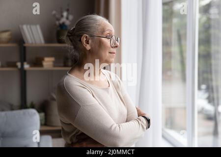 Donna più anziana in occhiali perso nei pensieri guardando fuori finestra Foto Stock