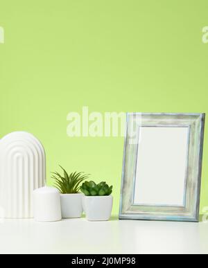 Cornice bianca vuota in legno e vasi da fiore con piante su tavola bianca, sfondo verde Foto Stock