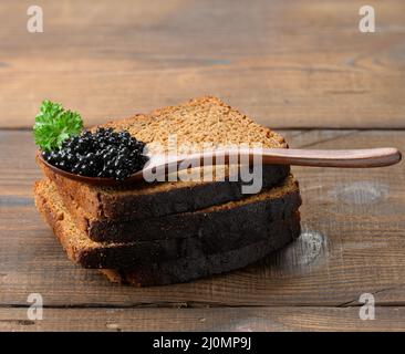 Caviale di paddlefish nero fresco e fette di pane in cucchiaio di legno marrone su un tavolo marrone, primo piano Foto Stock