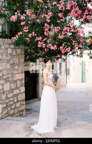 Sibenik, Croazia - 05.06.17: Sposa sorridente in un abito in pizzo bianco con un bouquet di lavanda si erge vicino ad un edificio in mattoni sotto a. Foto Stock