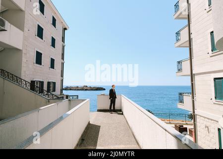 Uomo elegante in abito nero si erge su una terrazza tra due edifici sopra il mare e guarda in lontananza Foto Stock