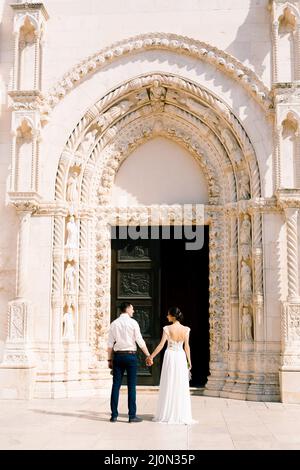 Sibenik, Croazia - 05.06.17: Sposare e sposare in un vestito di pizzo bianco reggendo le mani di fronte all'ingresso del Saint Jam Foto Stock