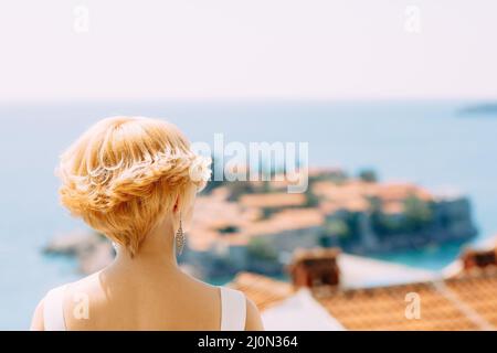 Sposa in un abito bianco si erge e guarda l'isola di Sveti Stefan vicino a Budva, Montenegro. Vista posteriore. Primo piano Foto Stock