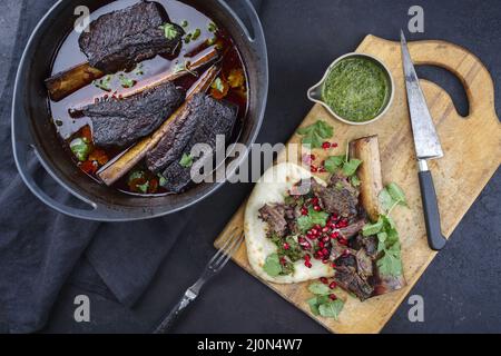 Traditional braised chuck beef ribs in red wine sauce and chimichurri served as top view in a modern design Dutch oven Stock Photo