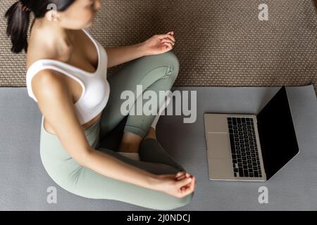 Scatto corto della donna asiatica meditating nel tappeto del pavimento a casa, ascoltando il podcast di meditazione sul laptop, pratica yoga cou in linea Foto Stock