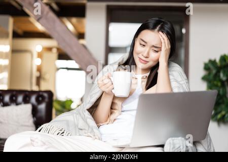 Giovane ragazza asiatica ammalata che rimane a casa in quarantena, cattura influenza o covid, guardando video su laptop e bere tè Foto Stock