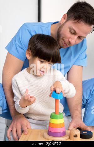 Giovane terapista che aiuta il ragazzo carino che ha paralisi cerebrale, giocando con lo sviluppo giocattolo in clinica di riabilitazione. Foto Stock