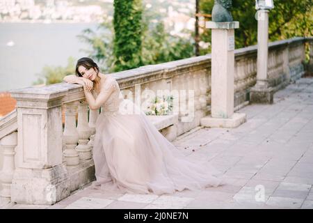 Sposa siede con la testa appoggiata su una balaustra di pietra in un giardino sulla costa della baia Foto Stock