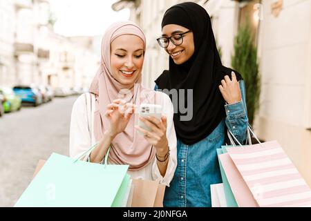 Due sorridenti donne islamiche di mezza età in foules che camminano all'esterno trasportando i sacchetti di shopping che guardano il telefono mobile Foto Stock
