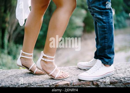 I piedi femminili in sandali accanto ai piedi maschili in sneakers si erigono su un bordo di pietra nel parco Foto Stock