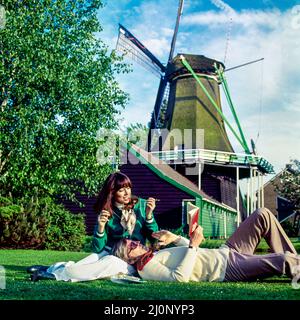 Vintage Netherlands 1970s, coppia di turisti di mezza età che si rilassano nel prato accanto ad un mulino a vento, Olanda, Europa, Foto Stock